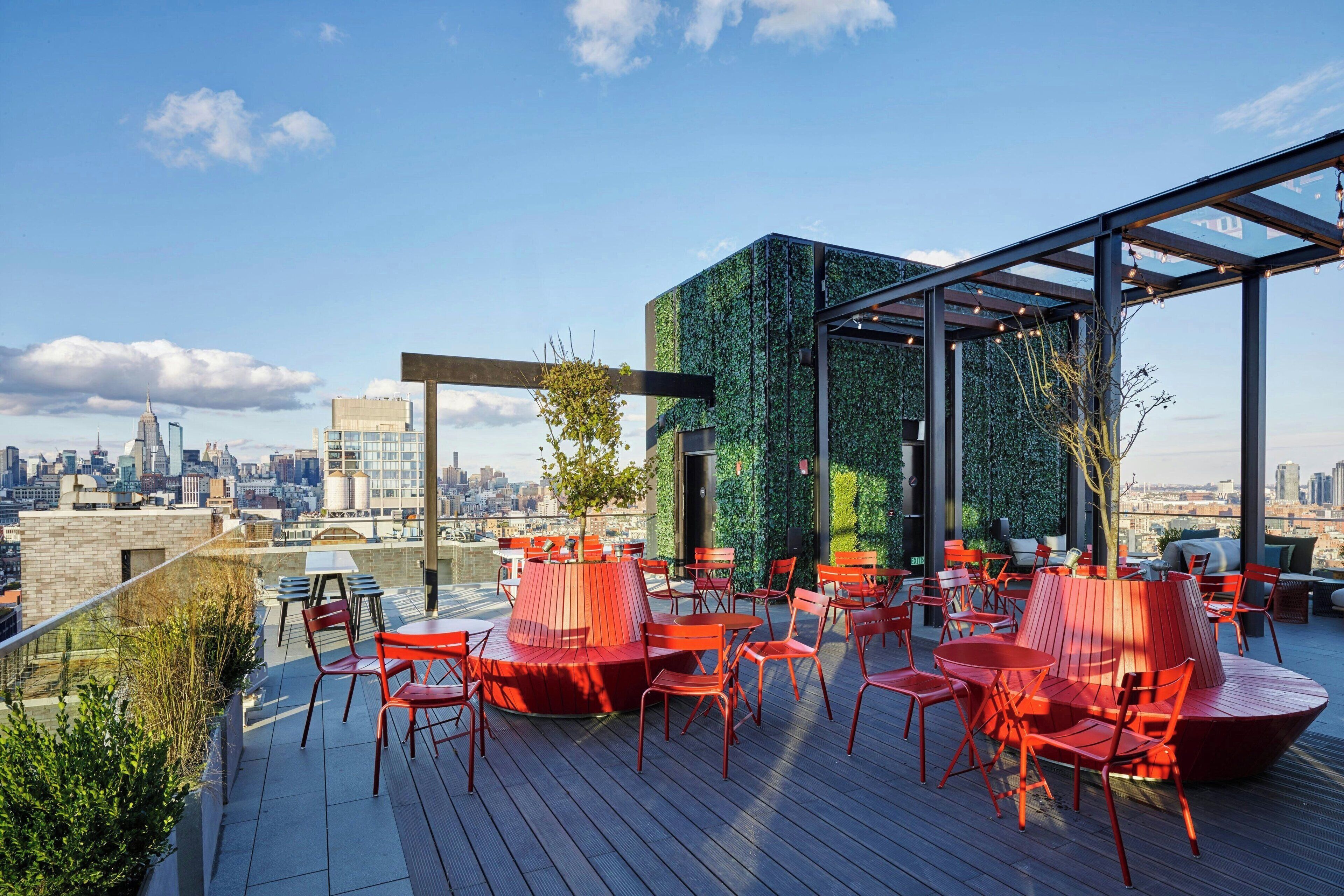 A rooftop terrace with red chairs and tables, a green wall, and a city view.