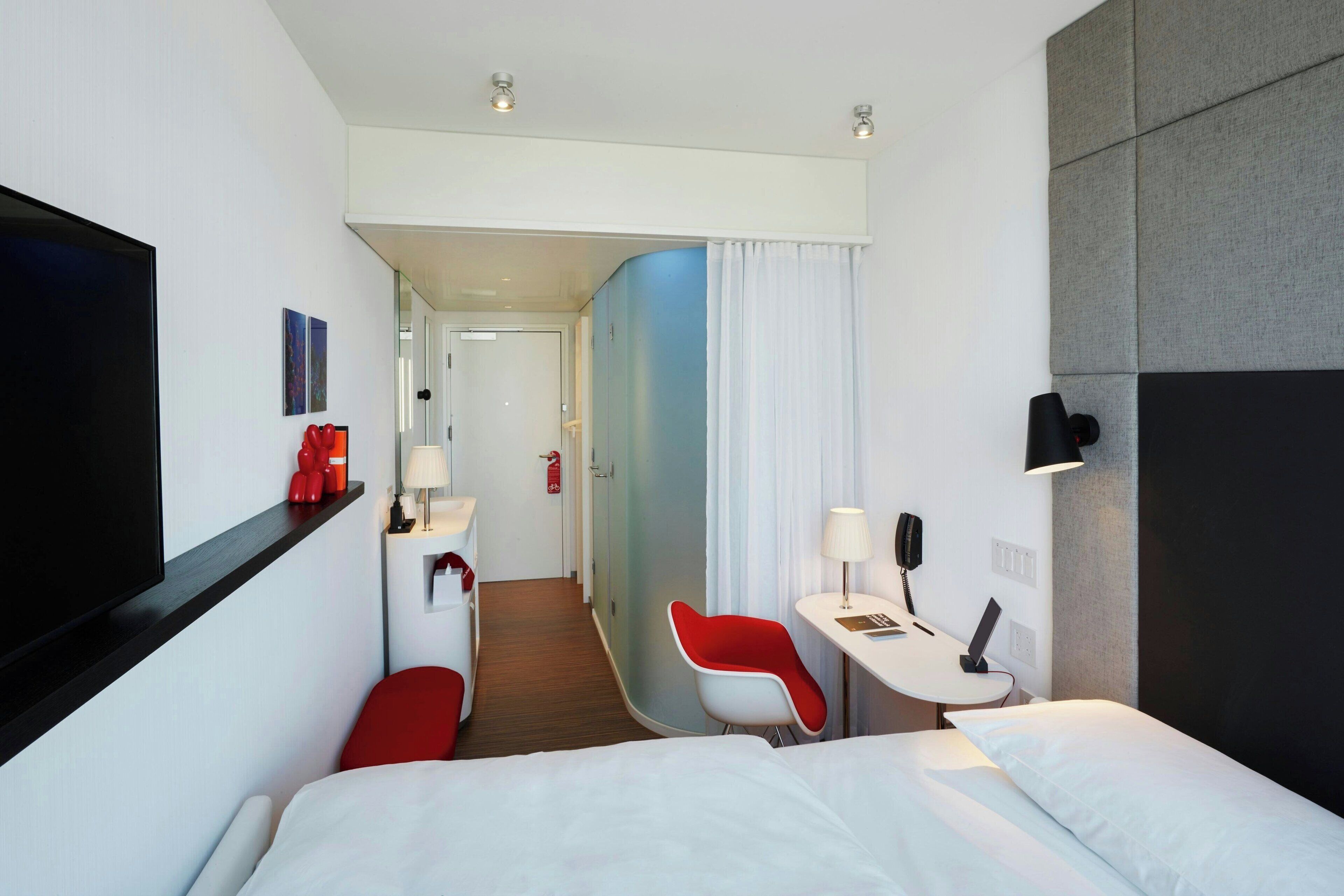 A hotel room with a bed, a desk, a chair, a TV, and a bathroom visible in the background.
