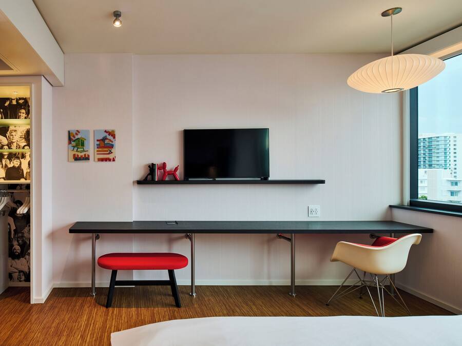 A modern room with a long desk, a red bench, a TV, and a window with a city view.