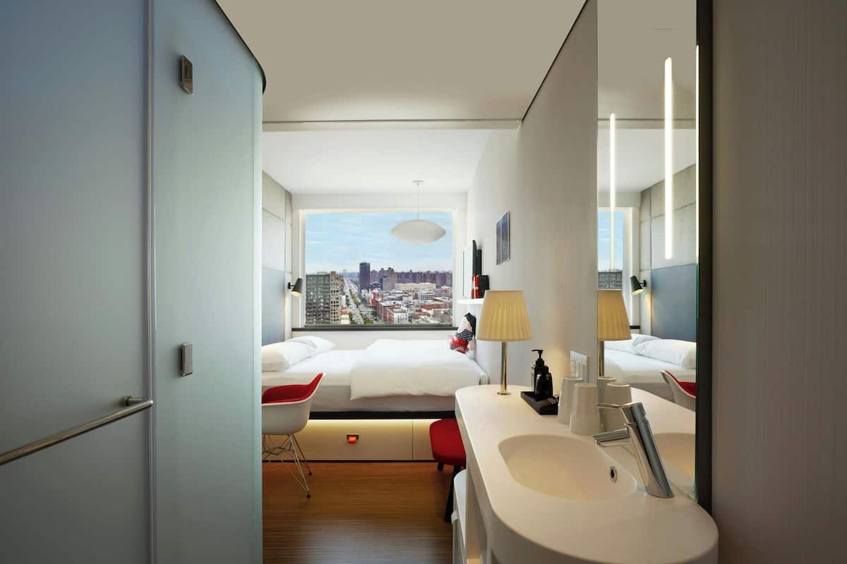A modern hotel room with a large bed, a bathroom with a sink and mirror, and a city view through the window.