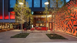 An outdoor seating area with tables and chairs, a building with a glass facade, and a wall with graffiti art. - citizenM New York Bowery (New York)