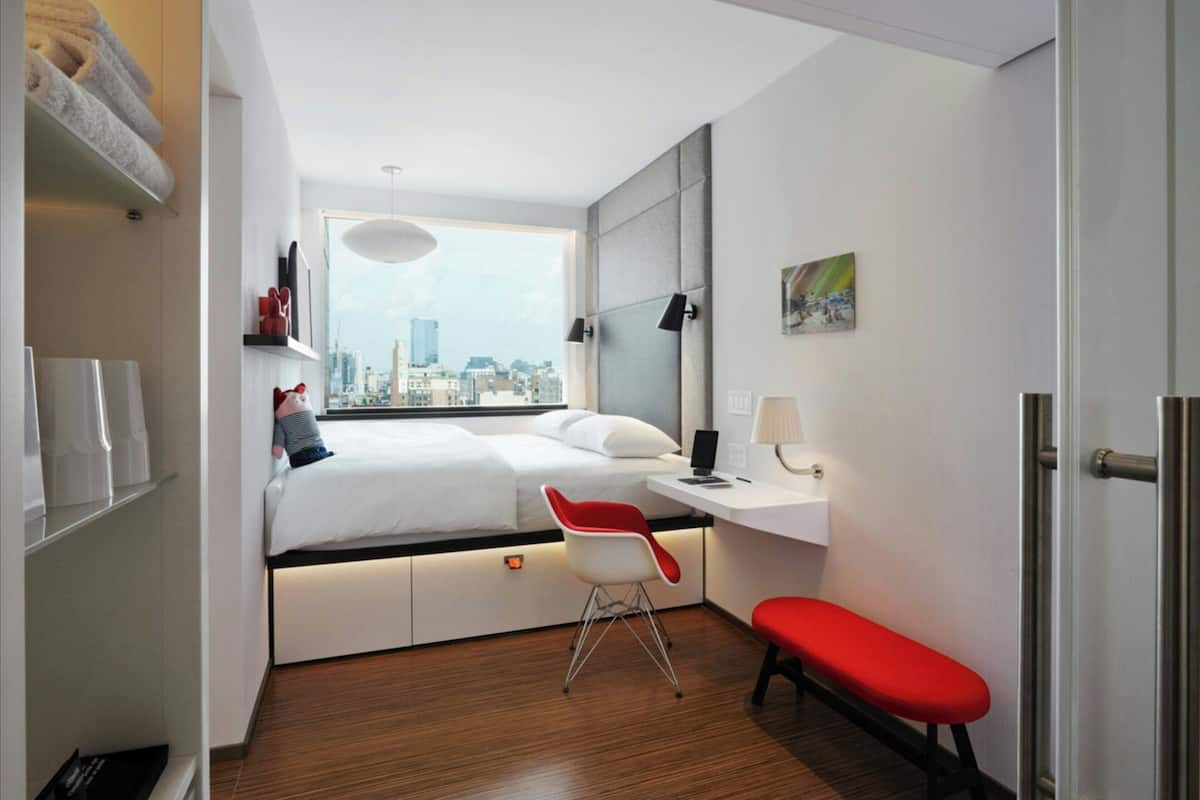 A modern hotel room with a bed, a desk, a red chair, and a view of the city.