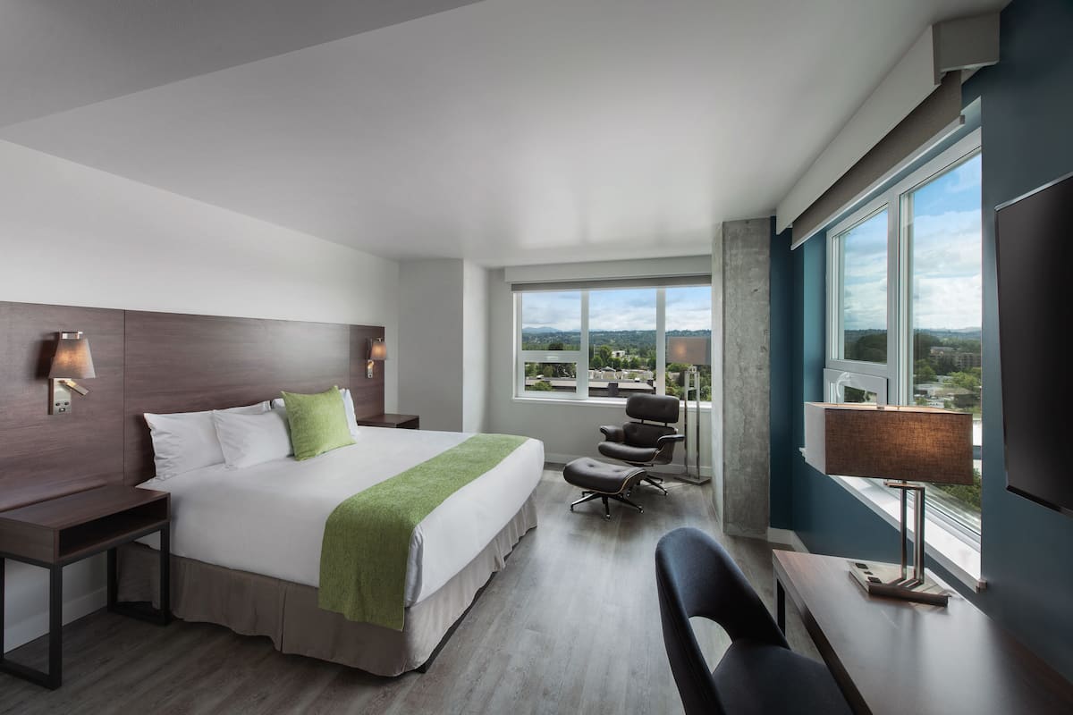 A modern hotel room with a large bed, a desk, a chair, and a view of the city through large windows.