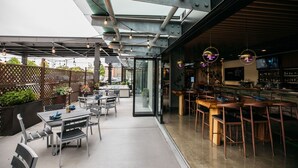 An outdoor dining area with tables and chairs, a bar area with stools, and a view of the street through a glass wall. - Hotel Interurban Seattle Airport SeaTac (Tukwila)