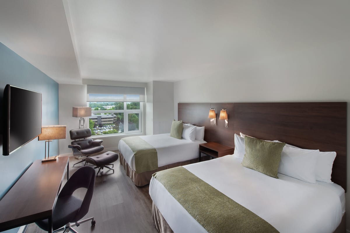 A hotel room with two beds, a desk with a chair, a TV, and a window with a view of buildings.