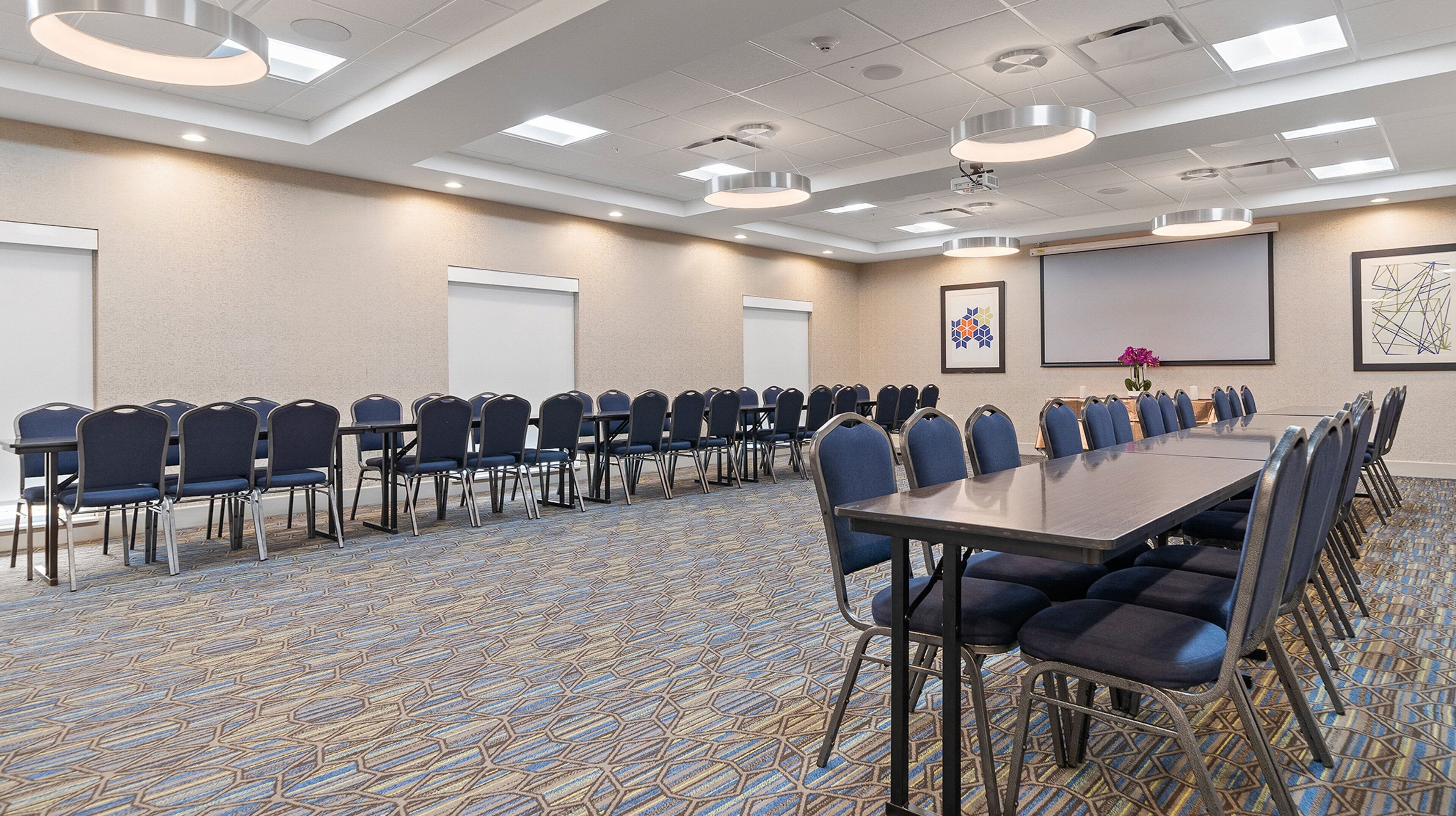 A conference room with a long table, rows of chairs, a projector screen, and a patterned carpet.