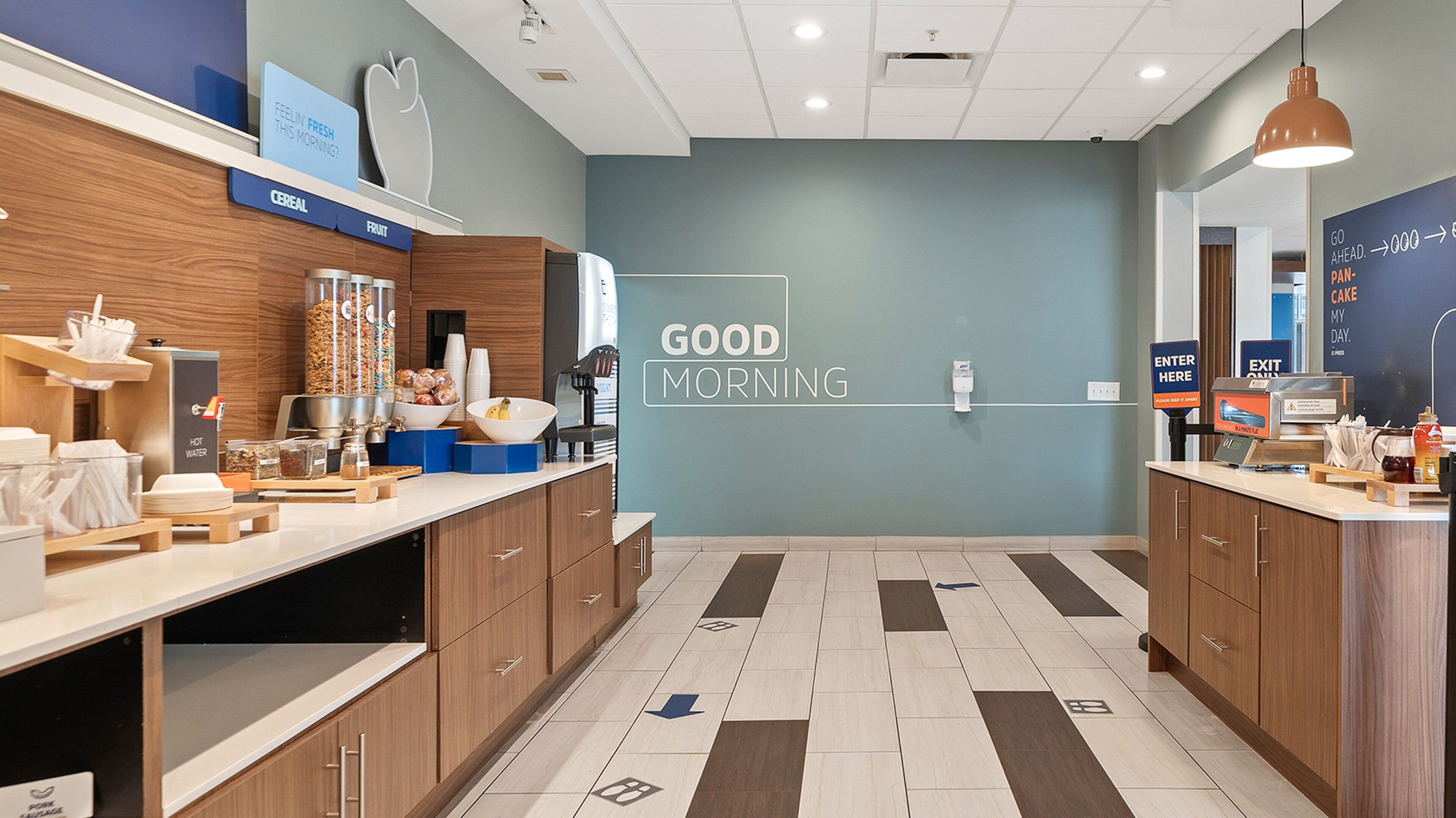 A modern breakfast area with a 'Good Morning' sign, a fruit and cereal display, and a water dispenser.