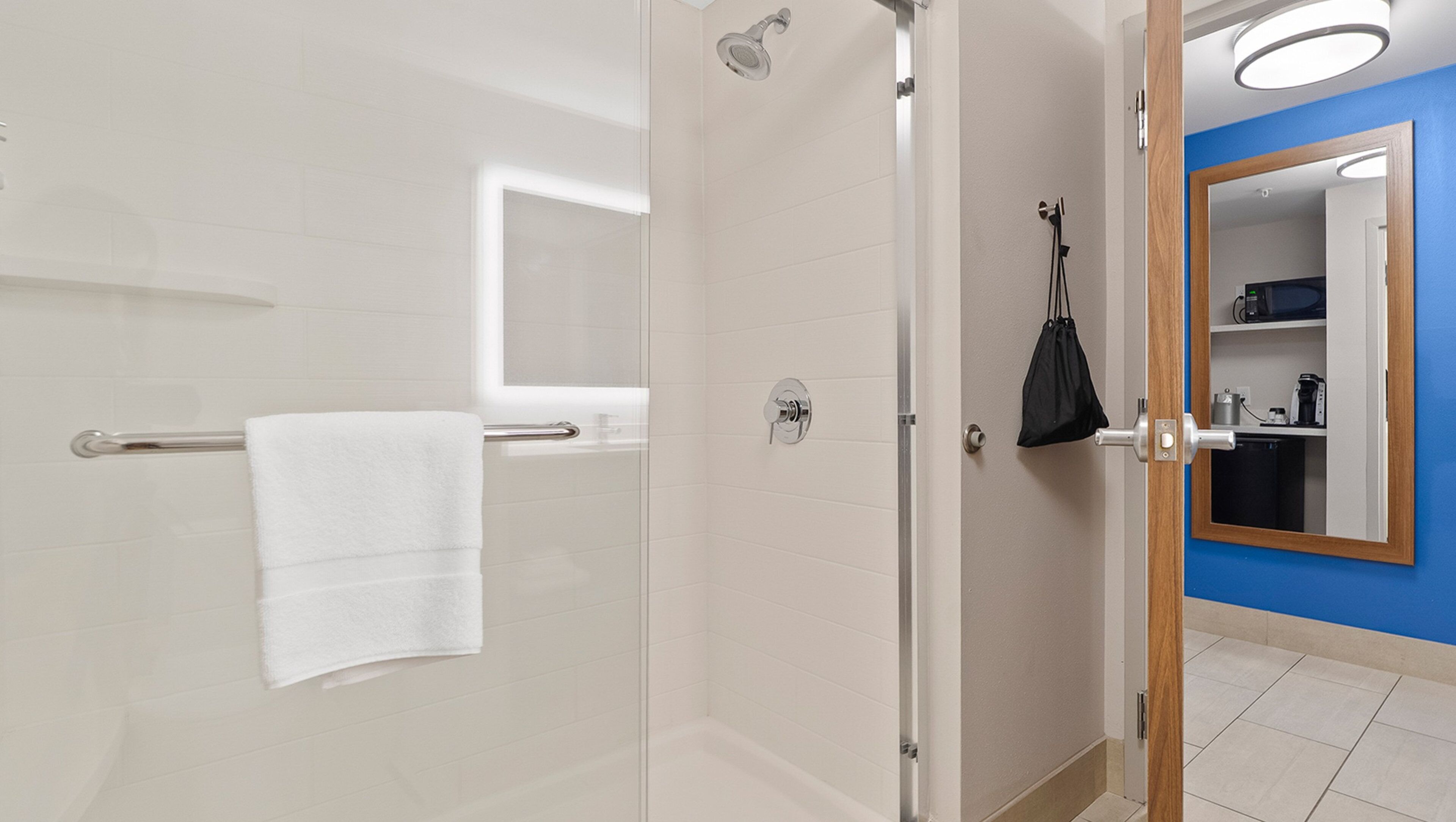 Un bagno moderno con cabina doccia in vetro, un asciugamano bianco su un porta asciugamani, una borsa nera appesa alla porta e una parete blu con uno specchio.