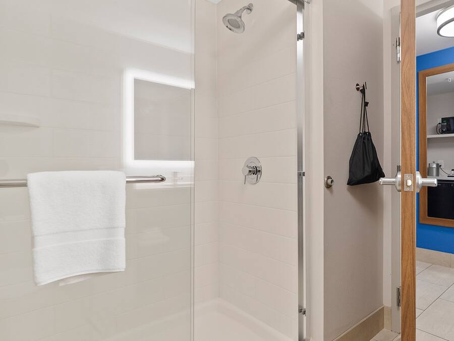 Un bagno moderno con cabina doccia in vetro, un asciugamano bianco su un porta asciugamani, una borsa nera appesa alla porta e una parete blu con uno specchio.