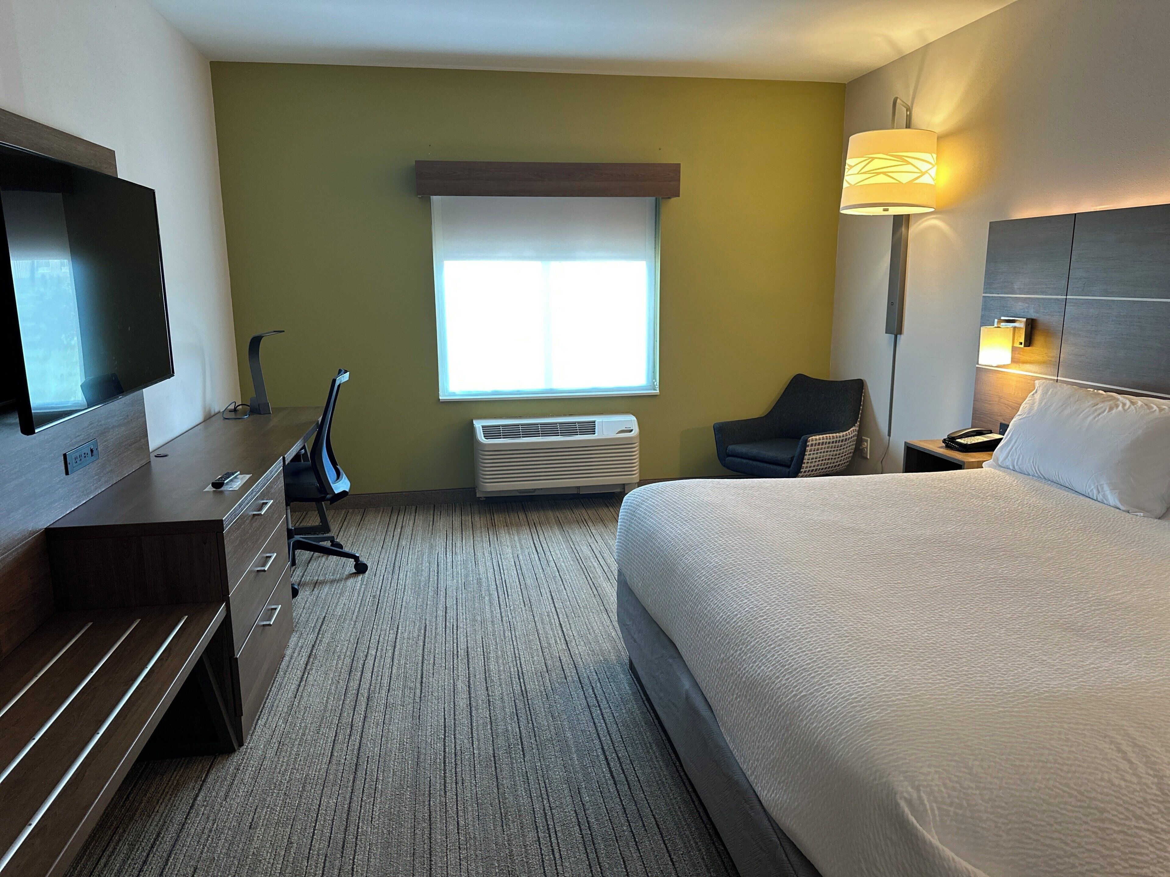 A hotel room with a bed, a desk with a TV, a chair, a bedside table with a lamp, and a window with blinds.