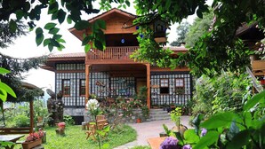Una casa de madera de dos pisos con un balcón, rodeada de vegetación exuberante y plantas en macetas.
