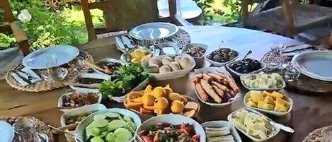 A round wooden table set with various food items and plates, surrounded by chairs, in an outdoor setting with greenery in the background.
