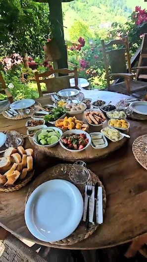 Una mesa redonda de madera dispuesta con diversos alimentos y platos, rodeada de sillas, en un entorno al aire libre con vegetación de fondo.