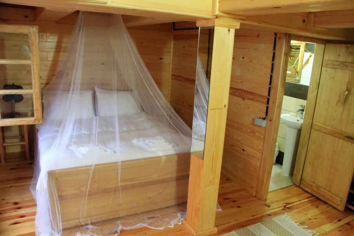 A wooden room with a bed under a canopy, a wooden cabinet, and a bathroom visible through an open door.