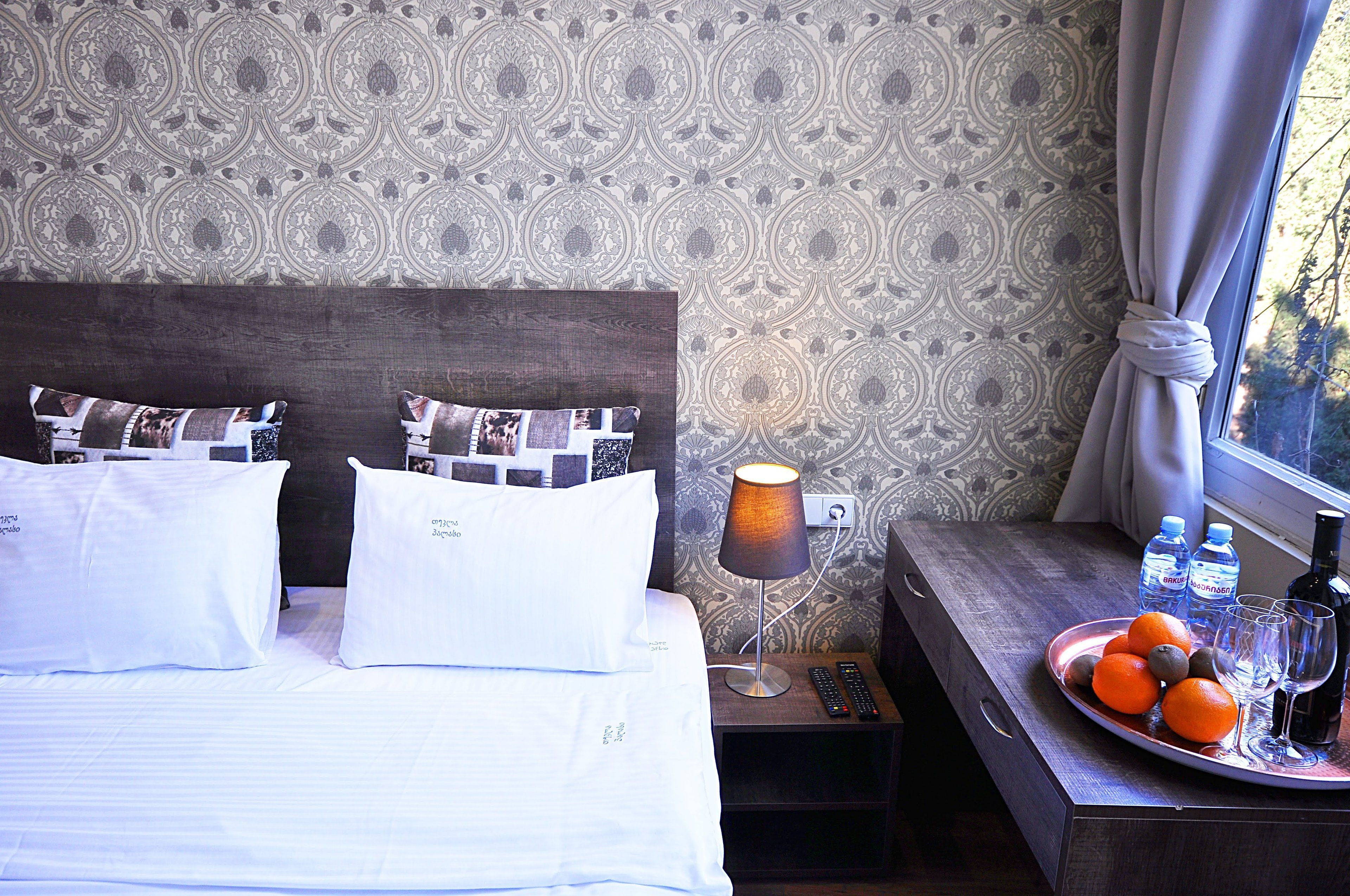 A neatly made bed with white linens, a bedside lamp, a tray with fruits and bottles, and a patterned wallpaper.