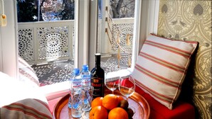 A tray with oranges, water bottles, a bottle of wine, and glasses on a red sofa by a window with patterned curtains.