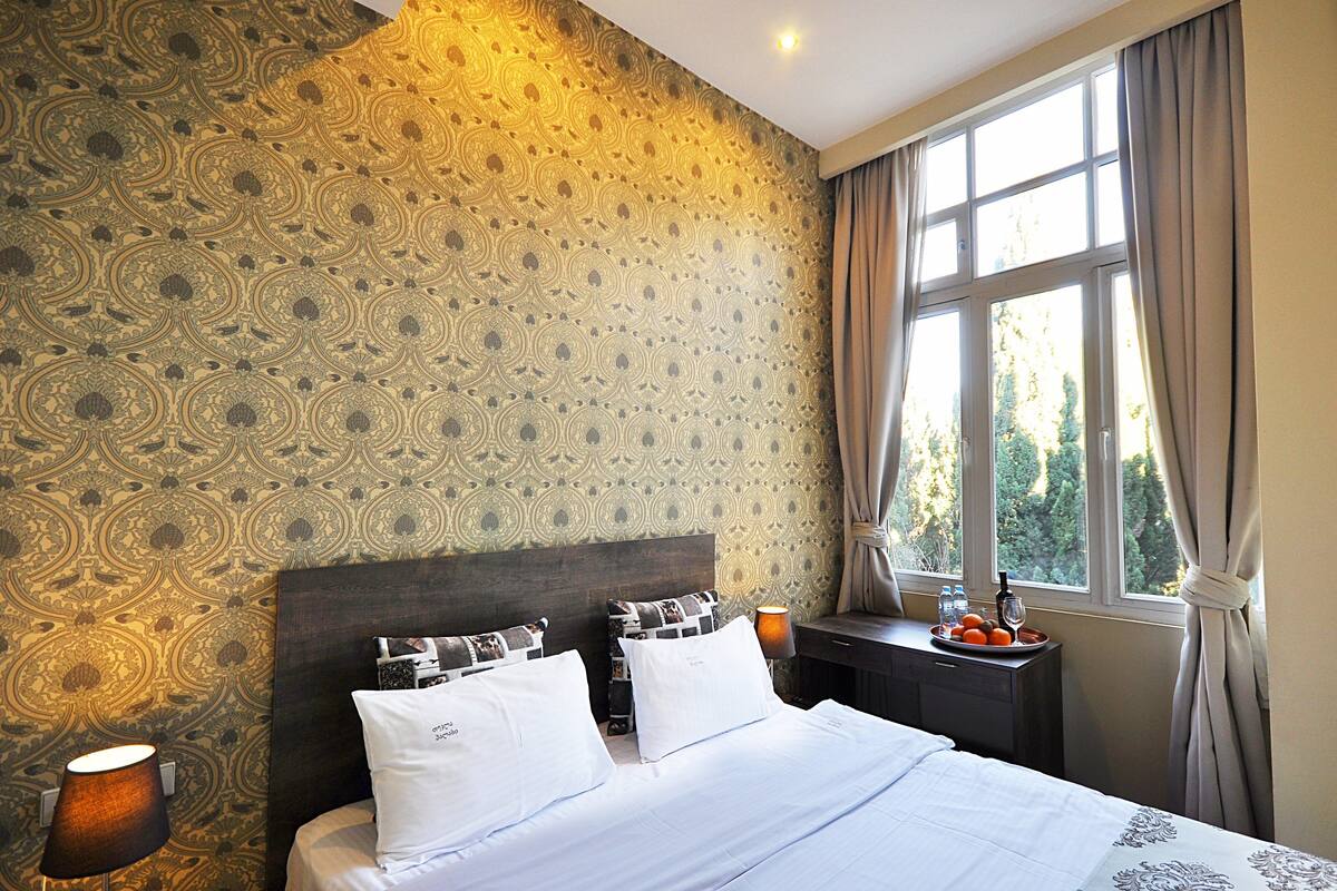 A hotel room with a patterned wallpaper, a large bed with white linens, a bedside table with a lamp, and a window with curtains.