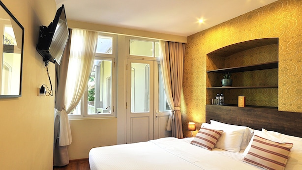 A hotel room with a bed, a TV mounted on the wall, a shelf with decorative items, and a window with curtains.