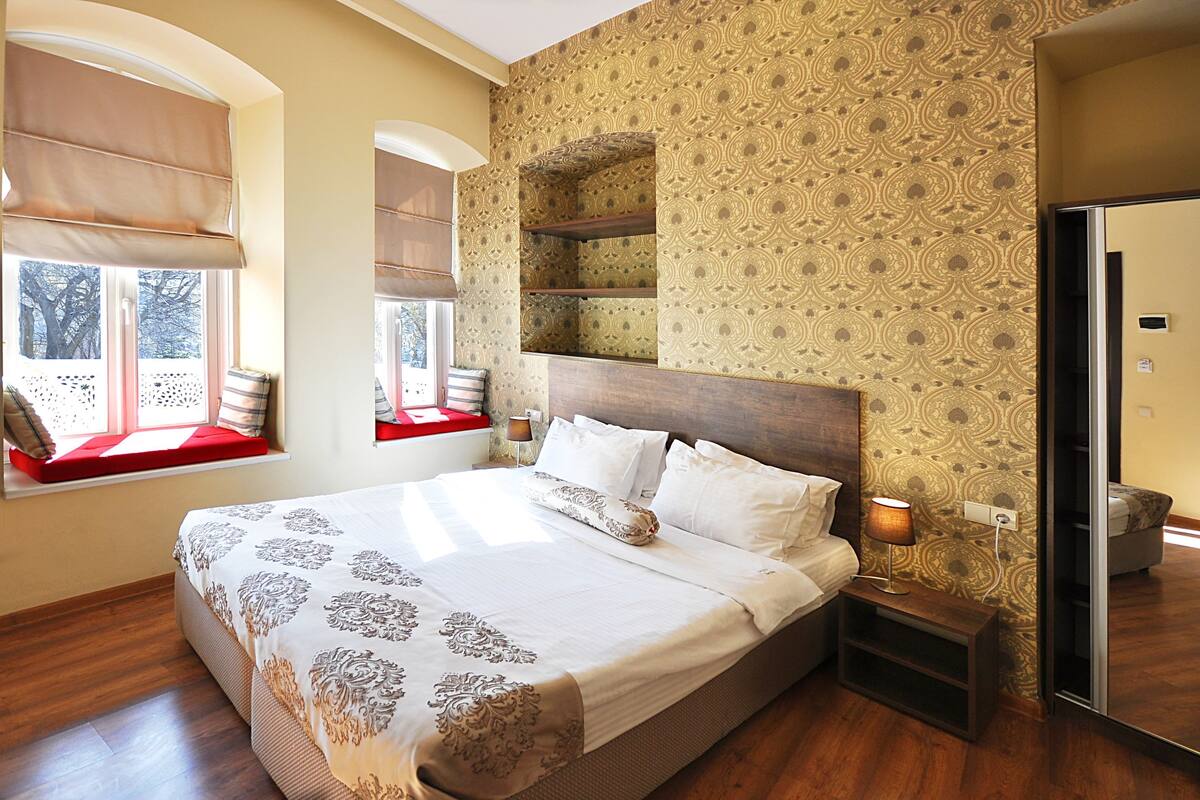 A neatly made bed with a patterned bedspread, a bedside table with a lamp, and a window with a view of trees.