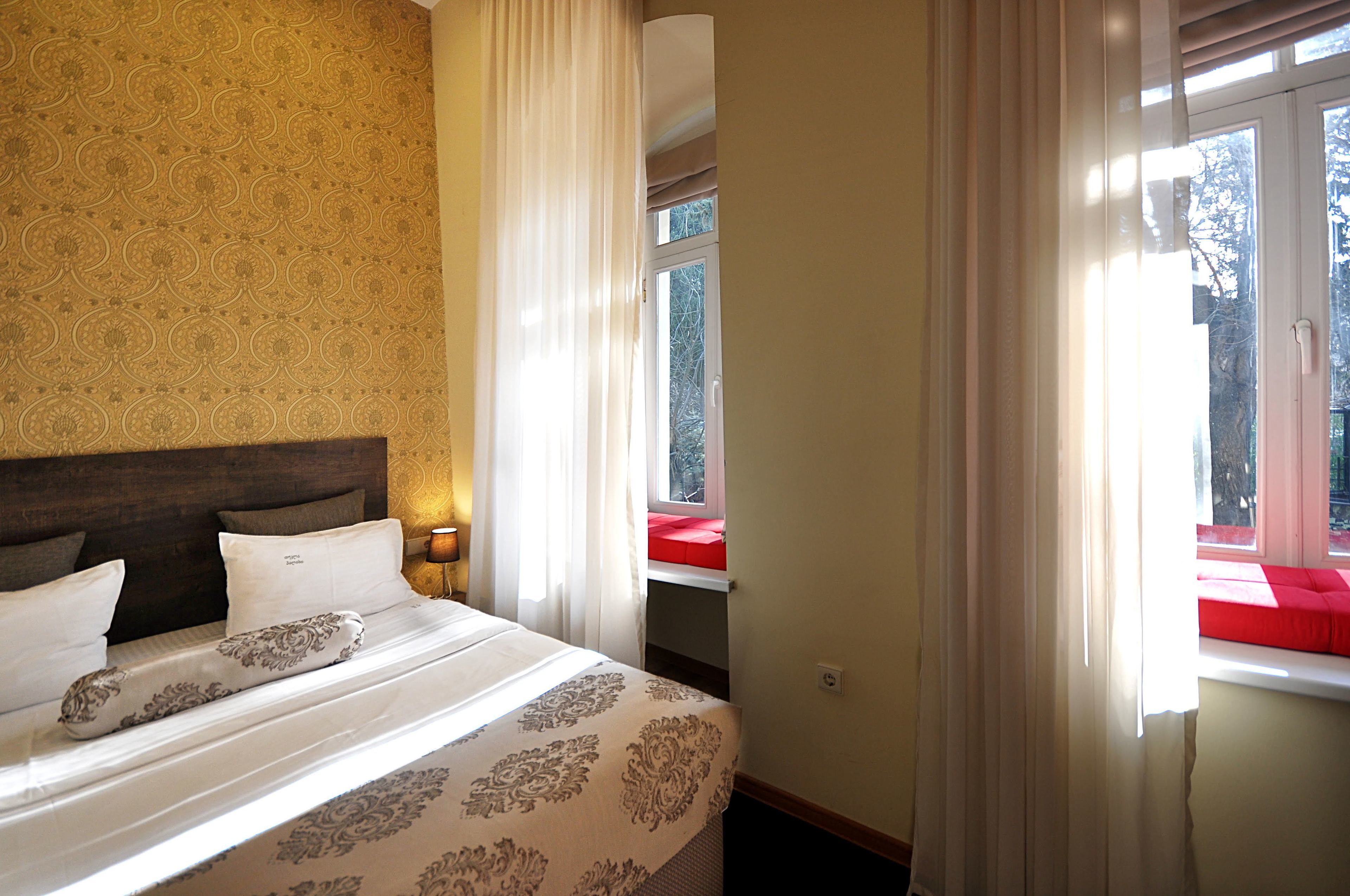 Habitación de hotel con pared estampada, cama con ropa blanca, mesita de noche con lámpara y ventana con asiento rojo.