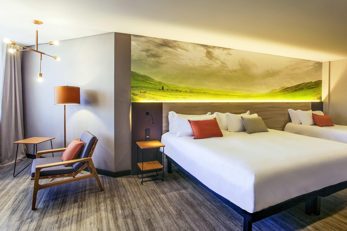 A hotel room with a large bed, a desk, a chair, and a wall-mounted artwork of a landscape.