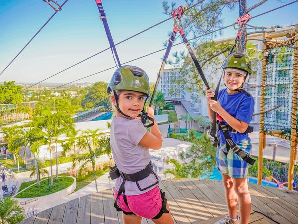 Two individuals on a zip line with safety harnesses, overlooking a resort area with palm trees and a pool.