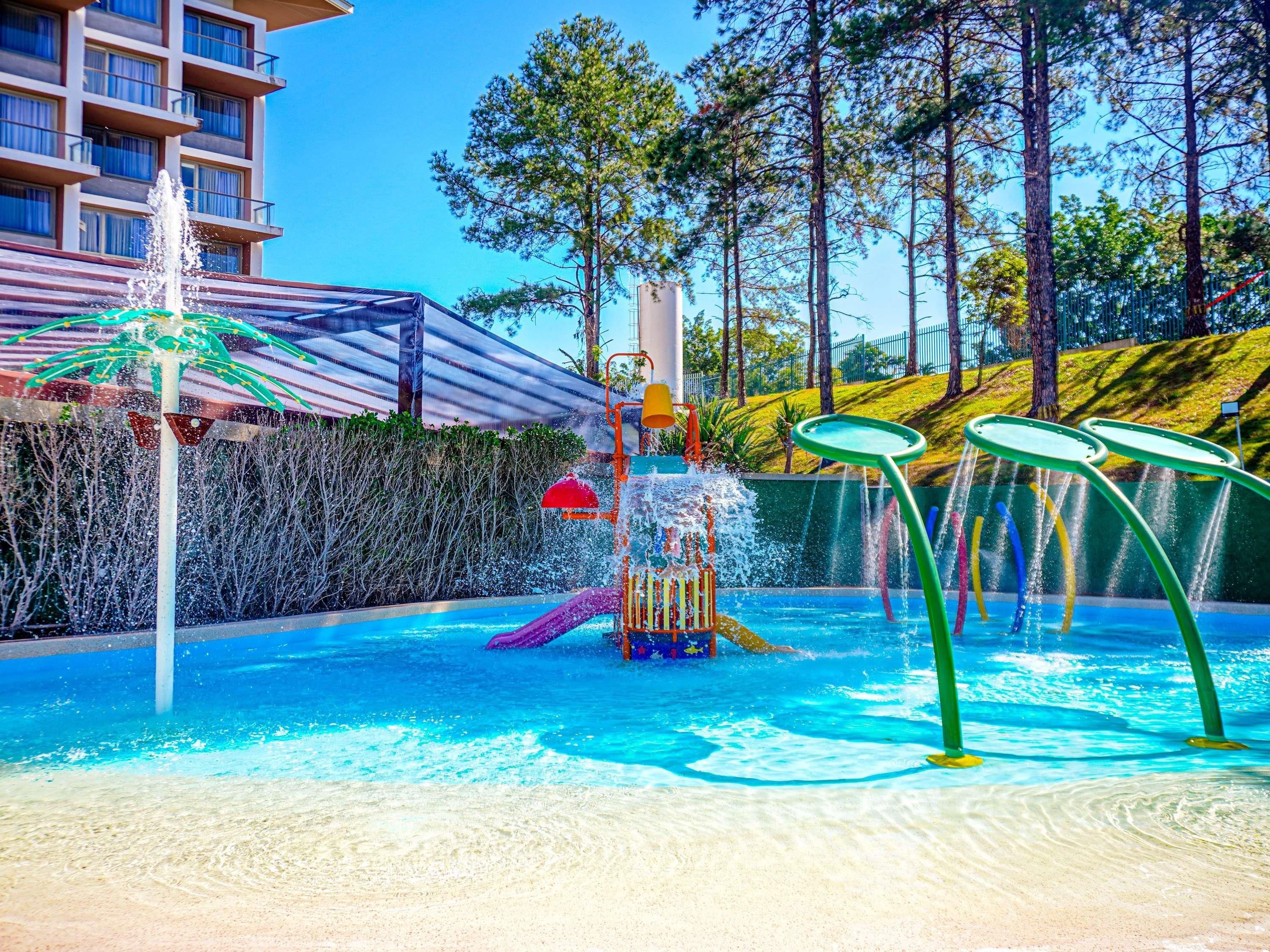 A swimming pool with a water play area, surrounded by a fence and trees.