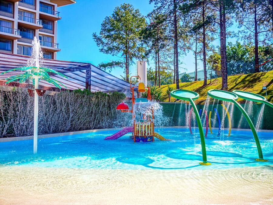 A swimming pool with a water play area, surrounded by a fence and trees.