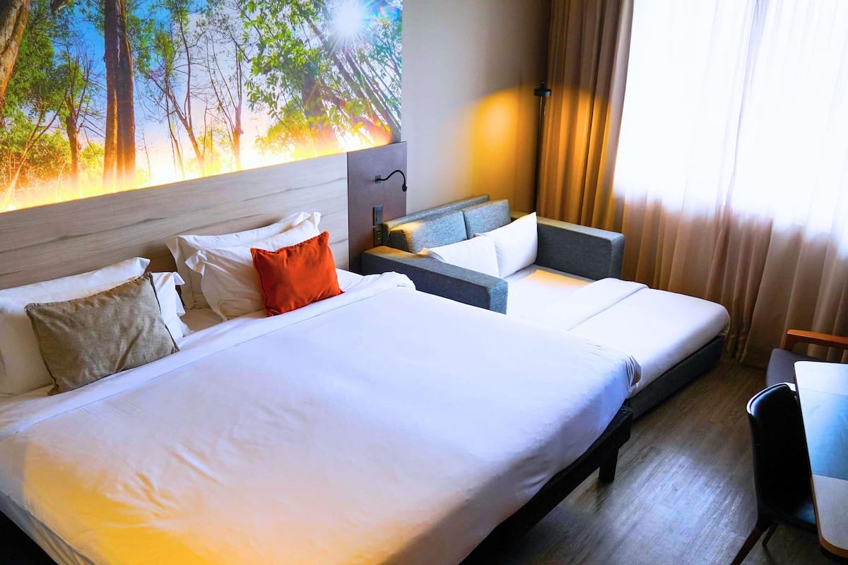 A hotel room with a large bed, a sofa, a desk, and a painting of a forest.