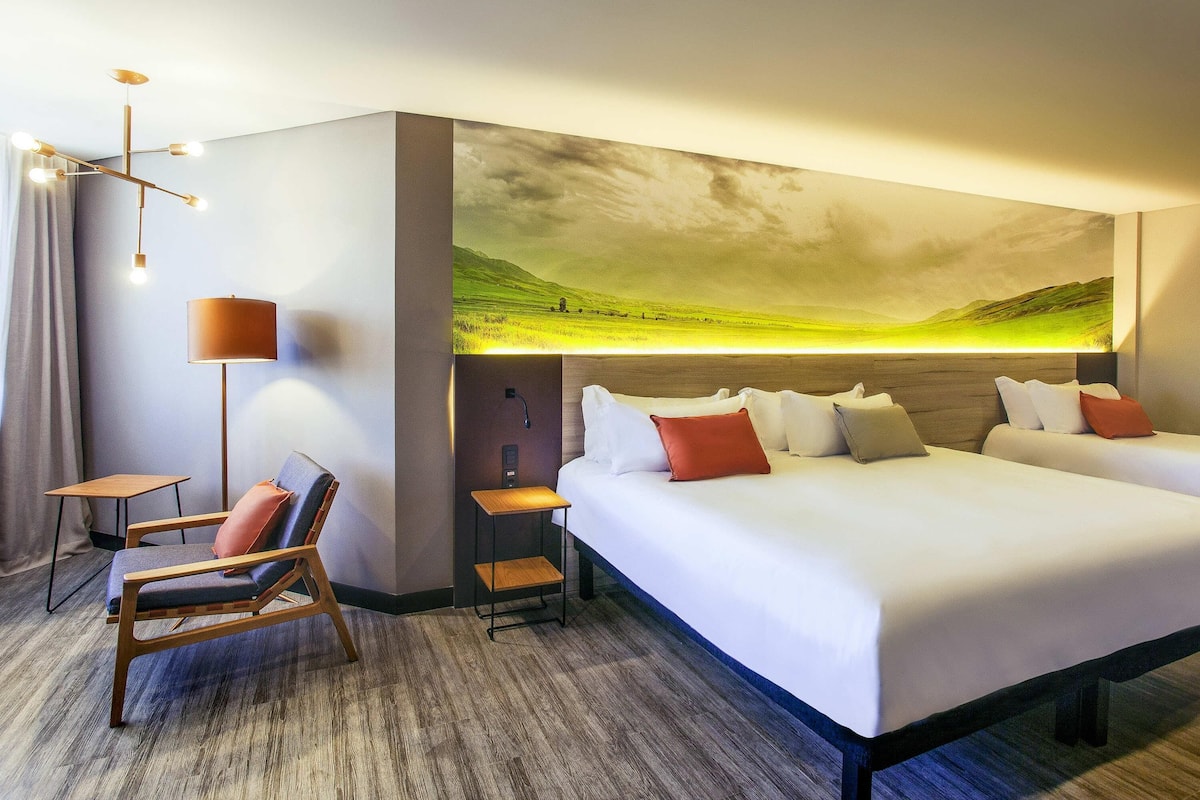 A hotel room with a large bed, a desk, a chair, and a wall-mounted artwork of a landscape.