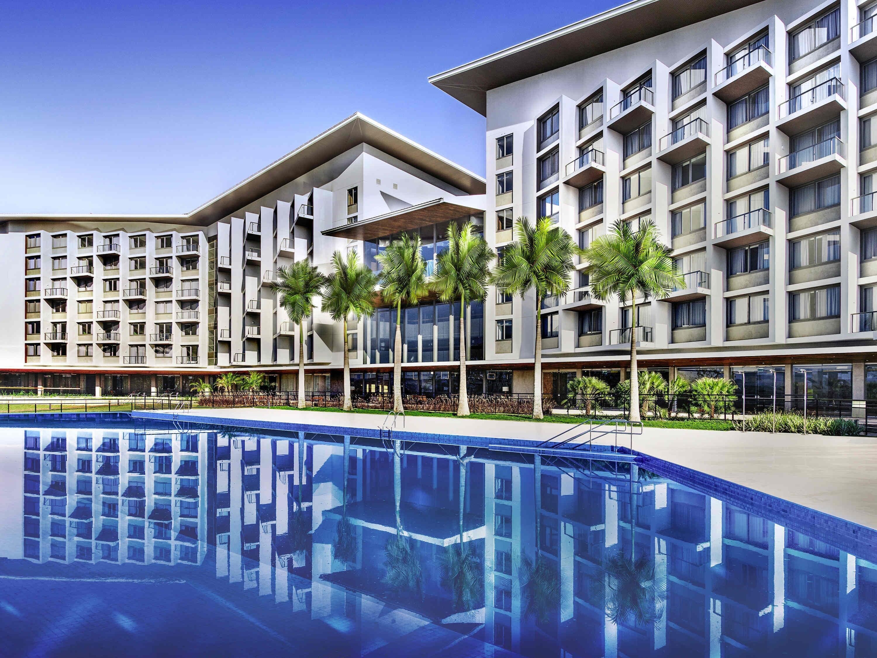 A modern hotel with a pool, multiple floors, balconies, and palm trees.