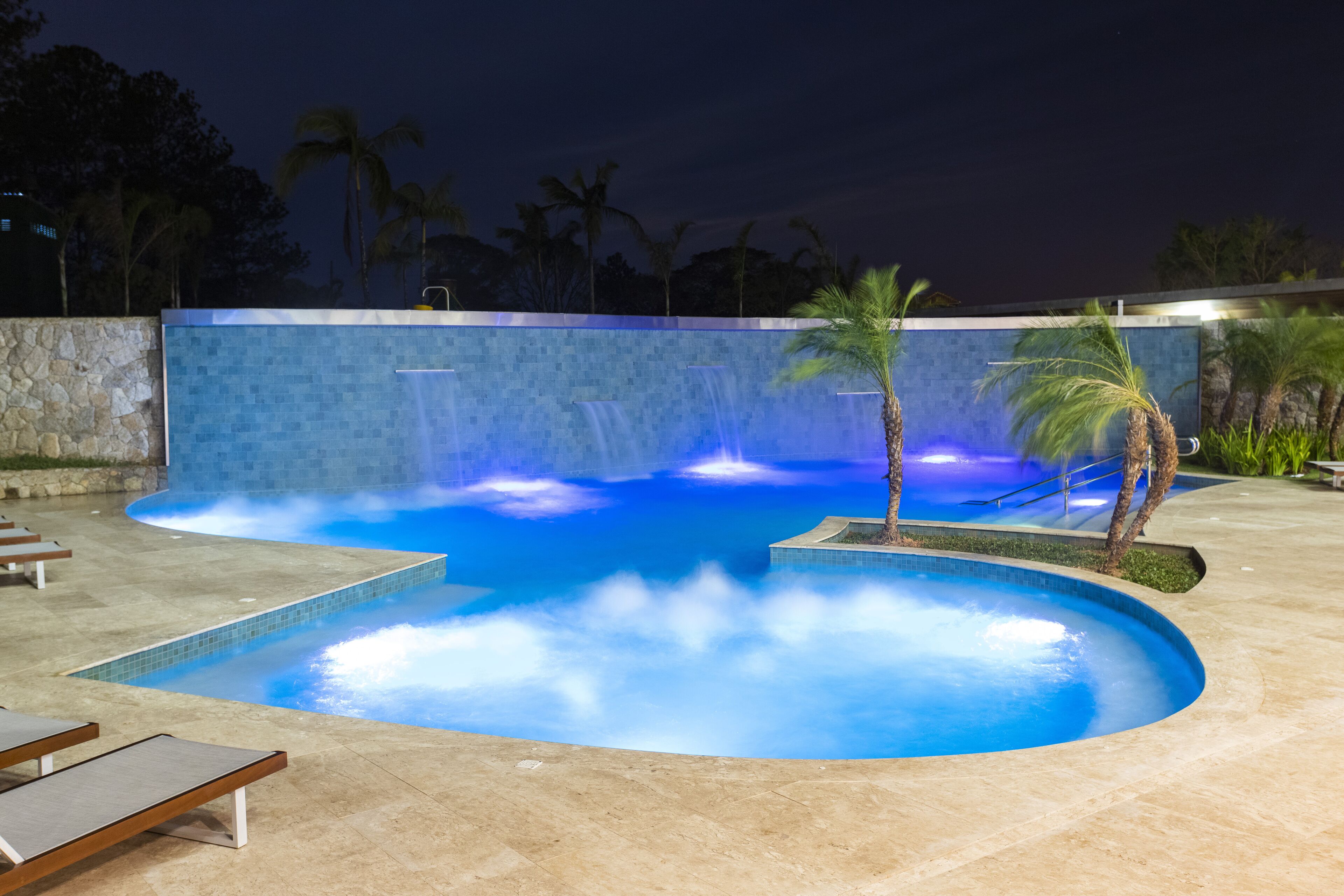 A swimming pool with a waterfall feature, surrounded by palm trees and lounge chairs.