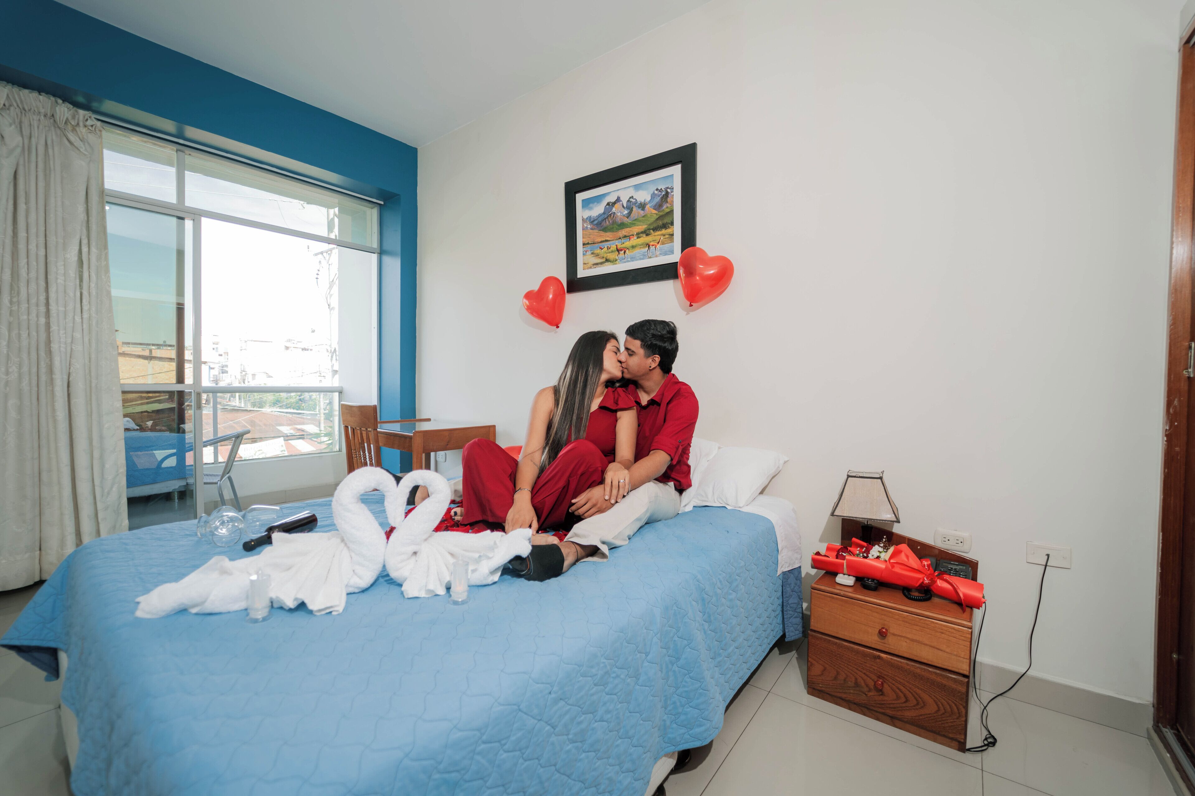 Habitación de hotel con cama, escritorio, silla, ventana con cortinas, un cuadro en la pared y cómoda con lámpara.