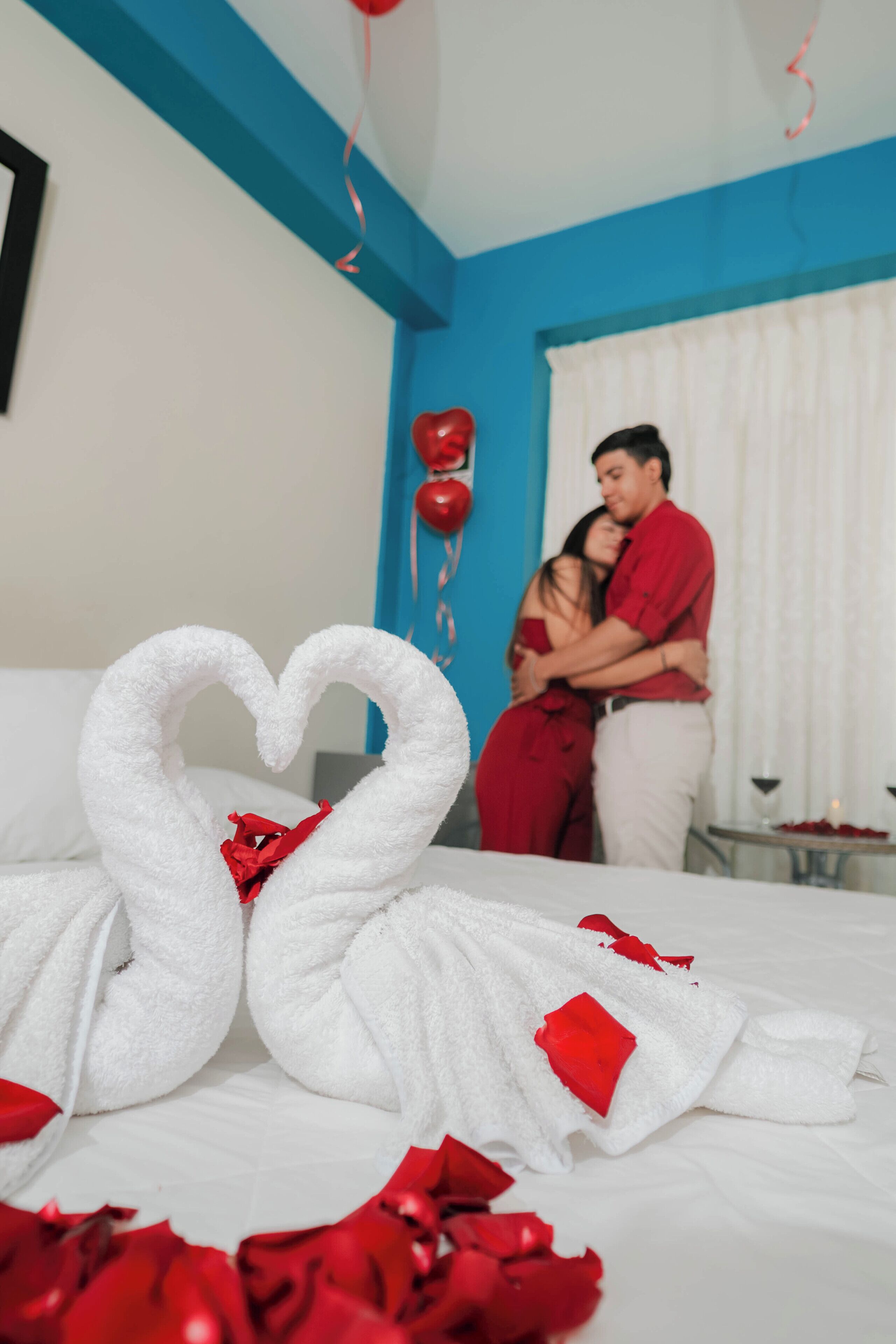 A hotel room with a bed, a heart-shaped towel arrangement, and a couple embracing.