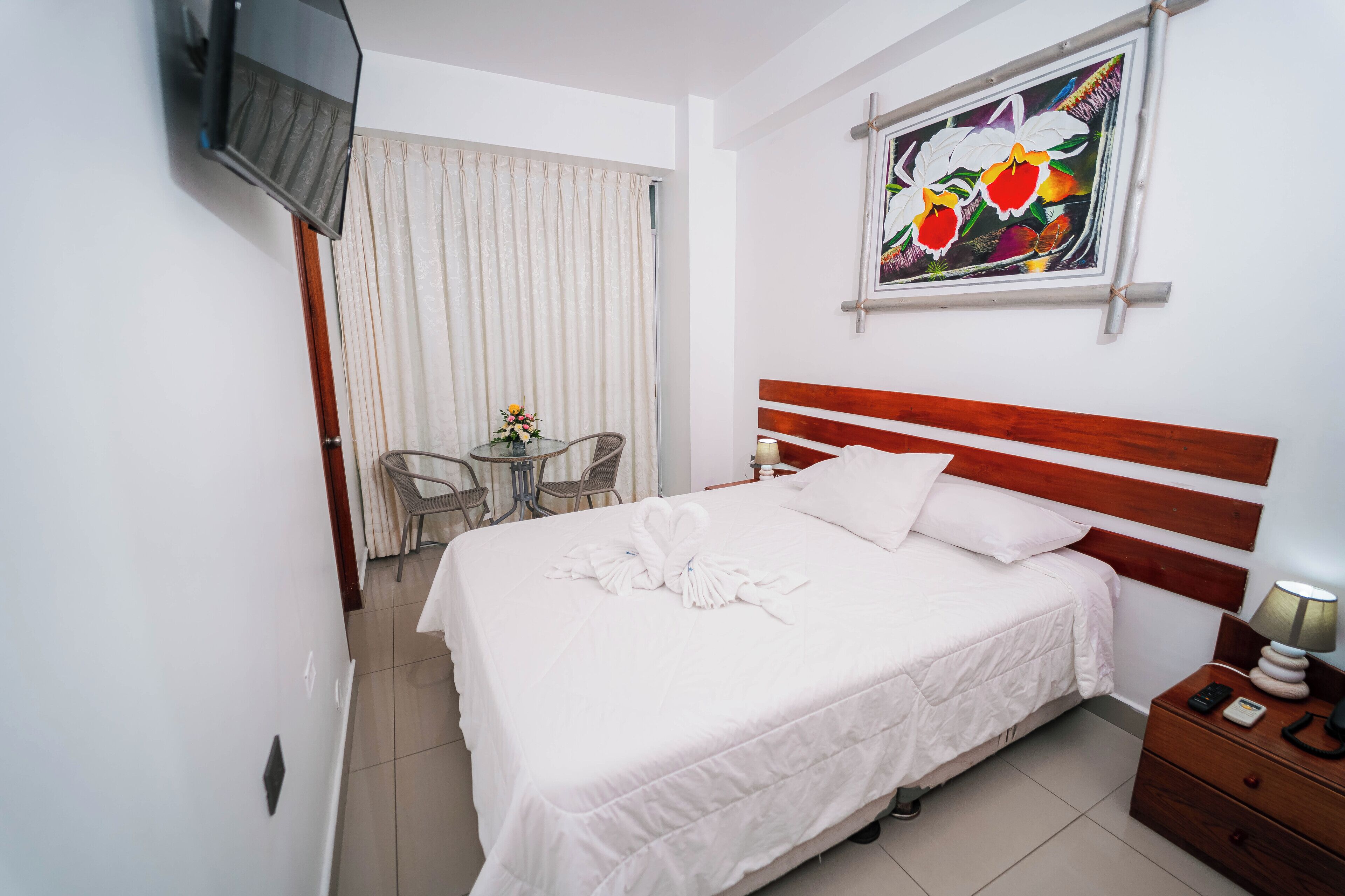 Habitación de hotel con una cama, un televisor en la pared, un cuadro, un escritorio con silla y una ventana con cortinas.