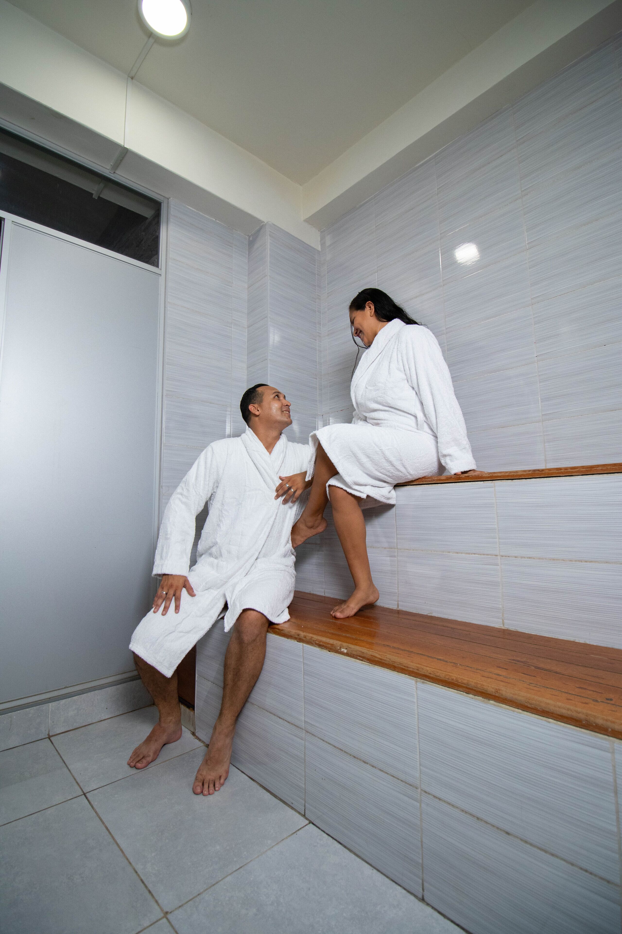 A bathroom with white tiled walls, a wooden bench, and a person sitting on the bench.