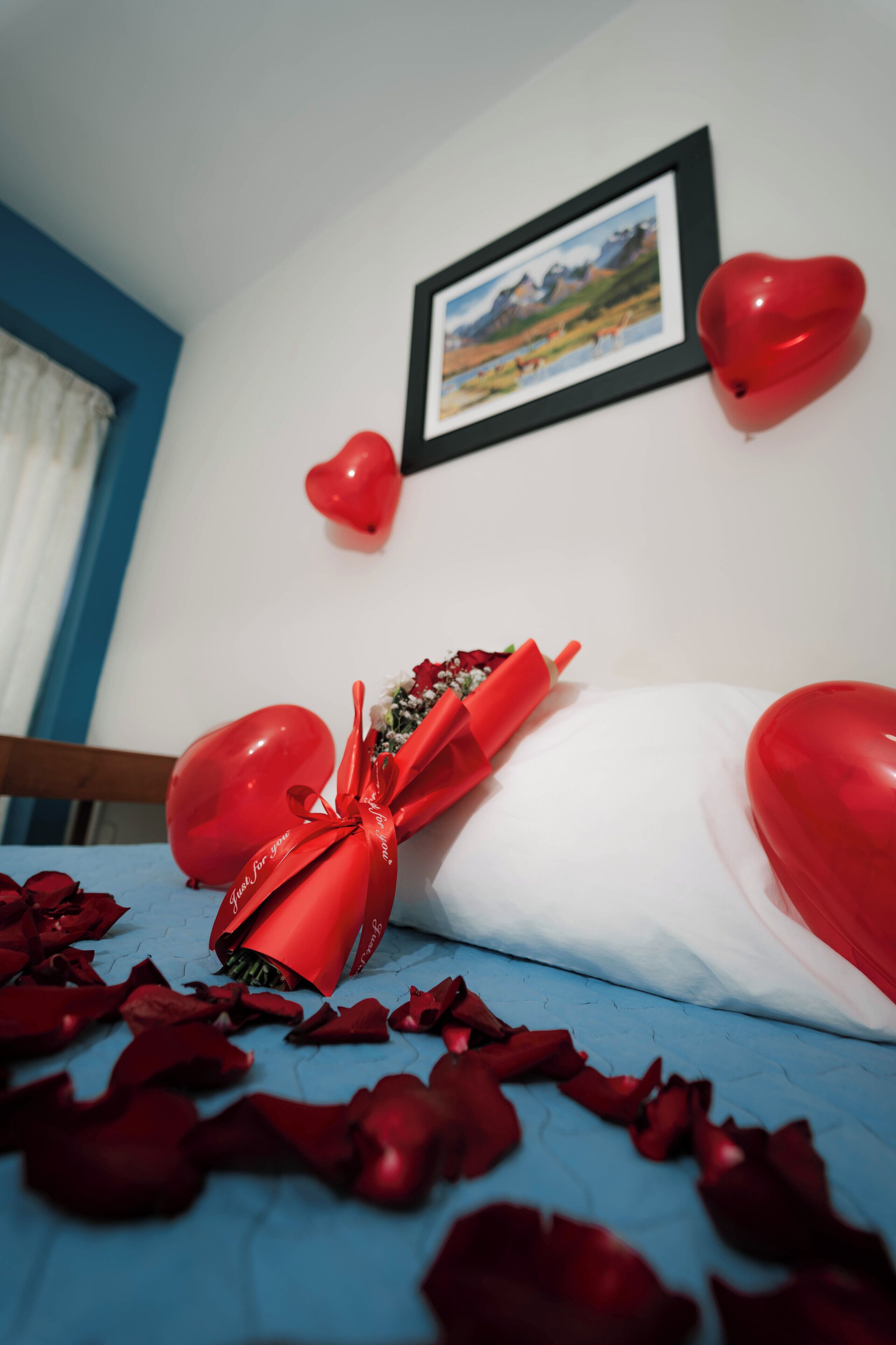 Una cama con pétalos de rosa roja esparcidos, un ramo de rosas rojas atado con una cinta roja y dos globos con forma de corazón rojo en la pared.
