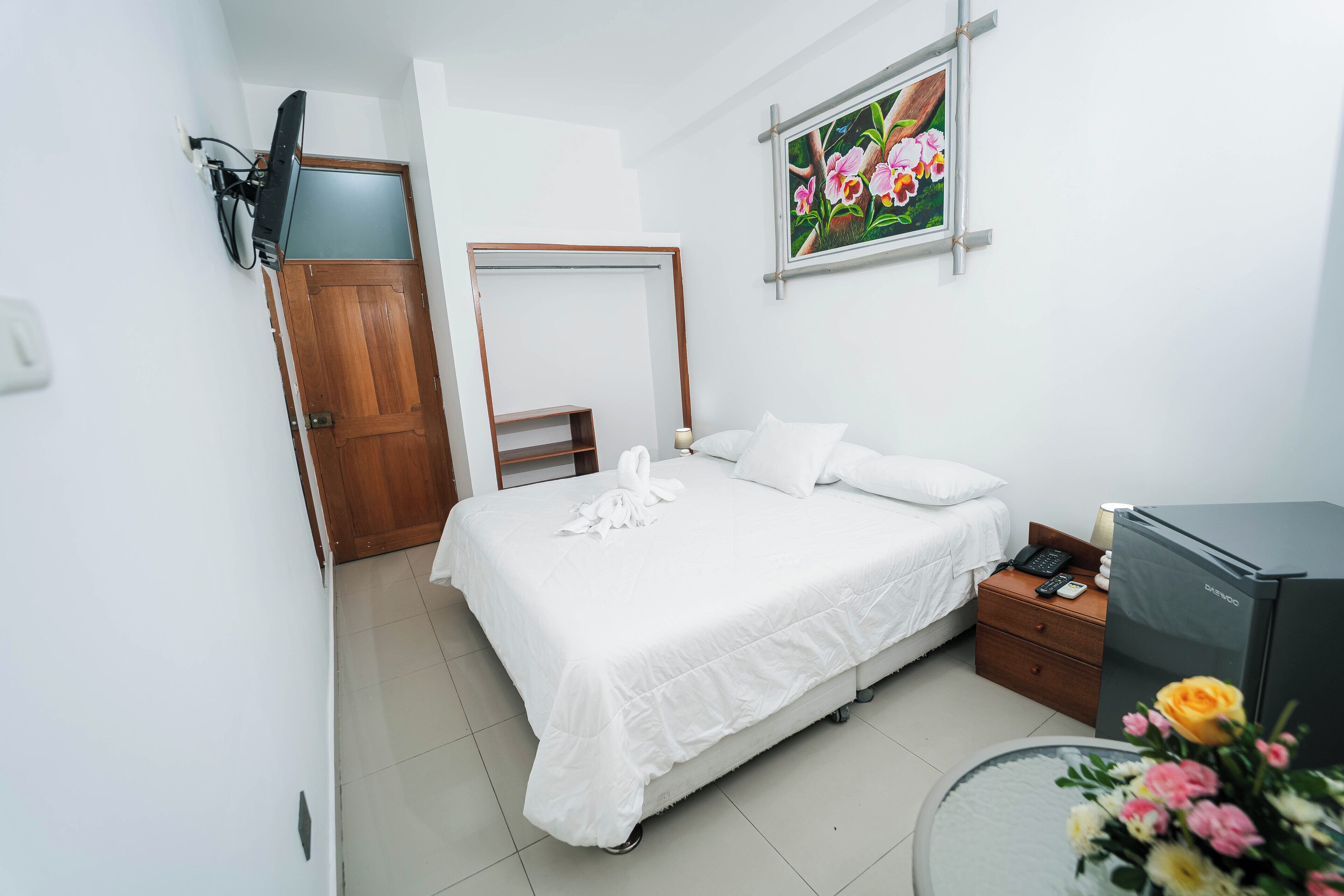 A hotel room with a bed, a bedside table with a lamp, a TV, and a painting on the wall.