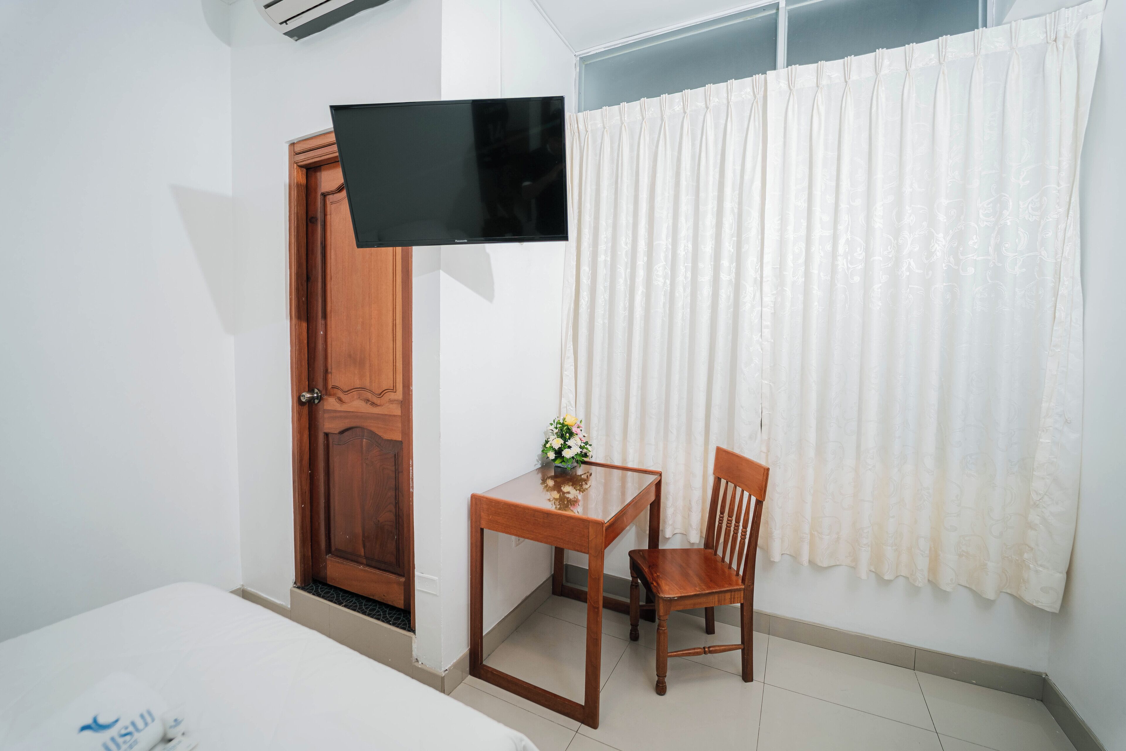 A hotel room with a bed, a wooden desk with a chair, a flat-screen TV mounted on the wall, and a window with white curtains.