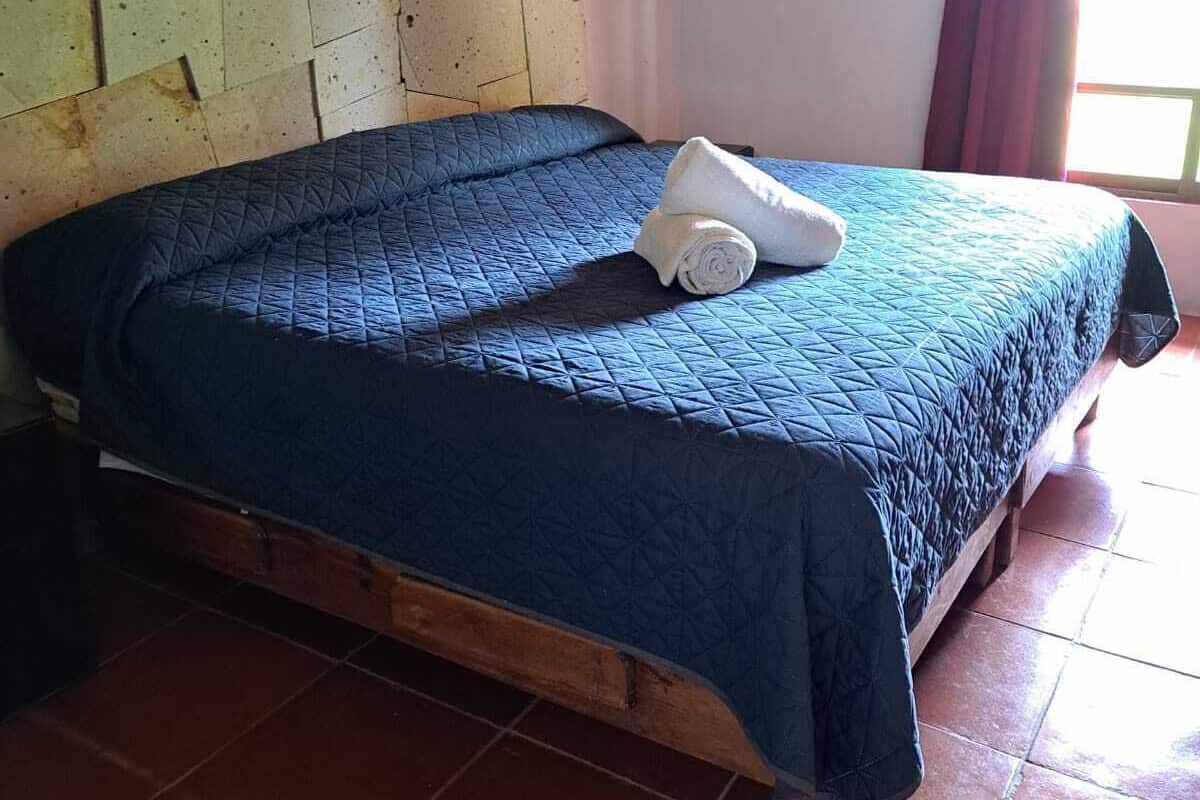 A single bed with a blue quilt, a rolled white towel, and a wooden headboard in a room with stone walls and tiled flooring.