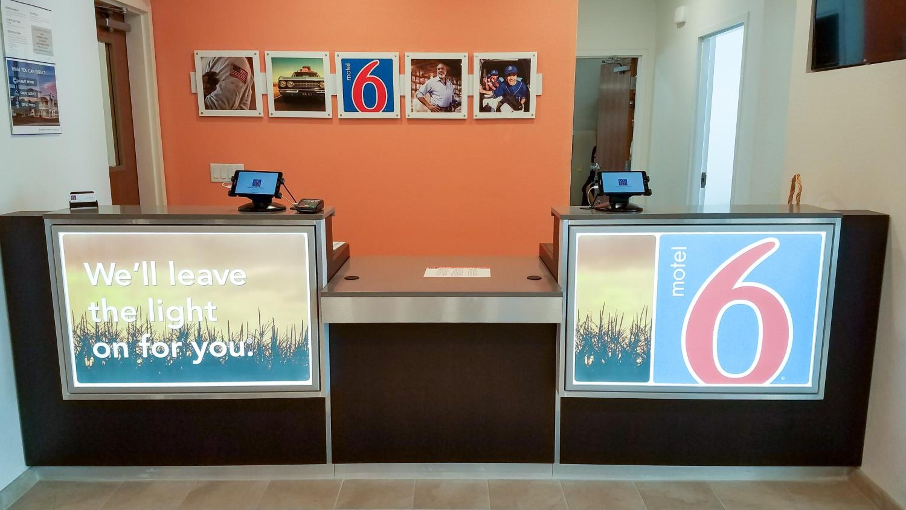 Taulell de recepció d'un motel amb dos monitors, un rètol de 'Motel 6' i un cartell que diu 'Deixarem la llum encèsa per tu'.