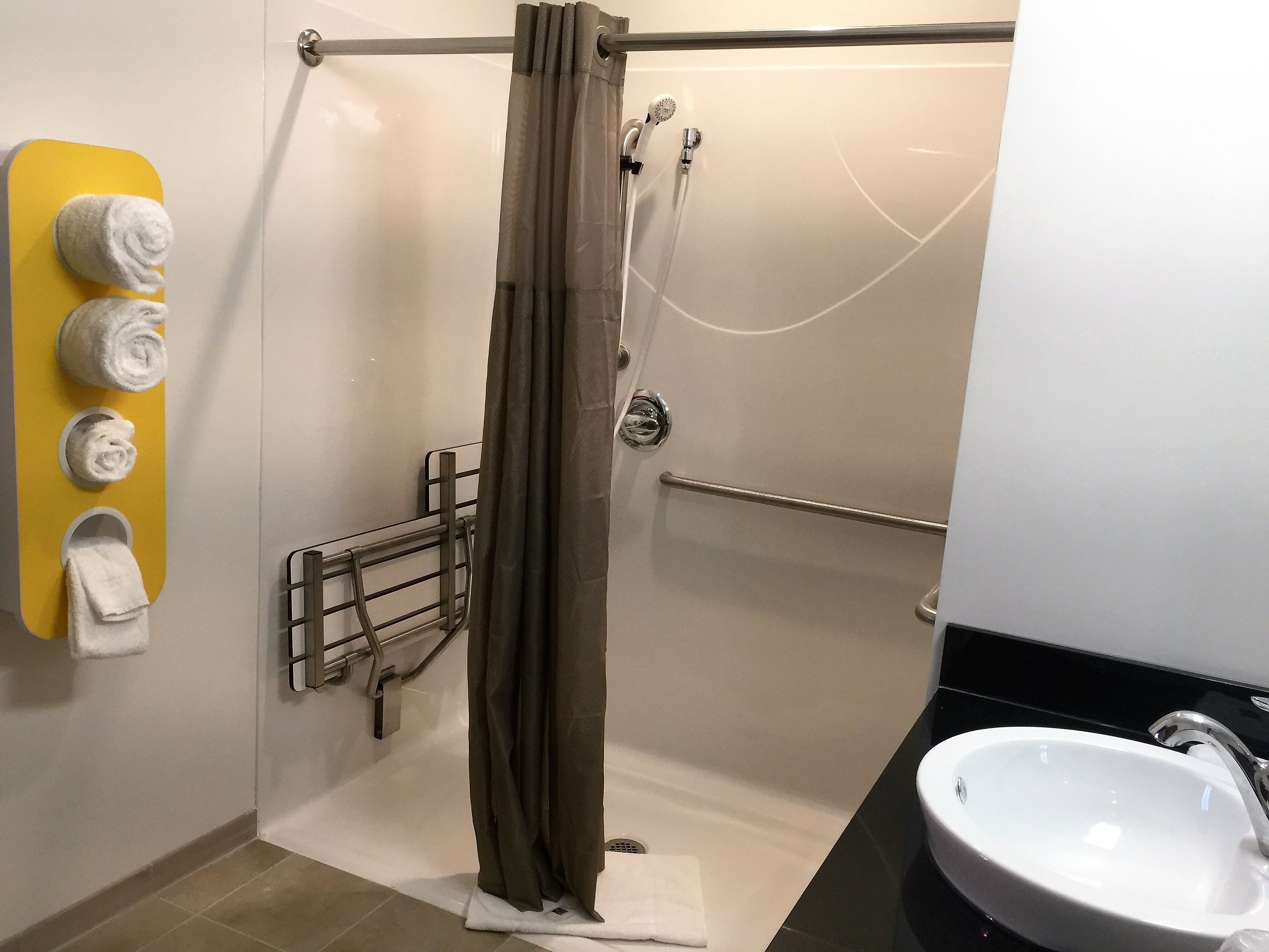A modern bathroom with a shower, a towel rack with three towels, a handrail, and a sink.