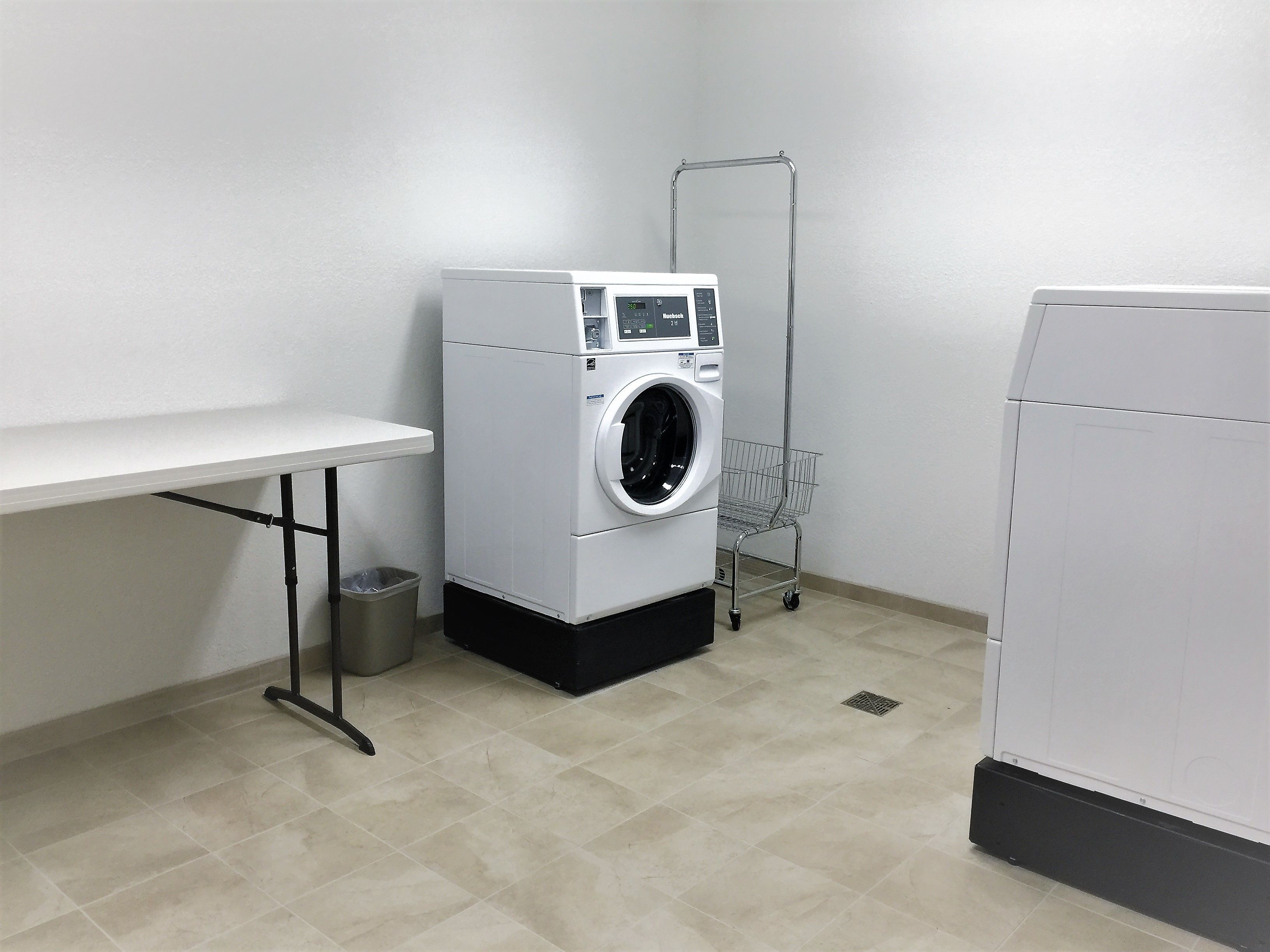 A laundry room with a washing machine, a dryer, a metal laundry cart, and a trash bin.