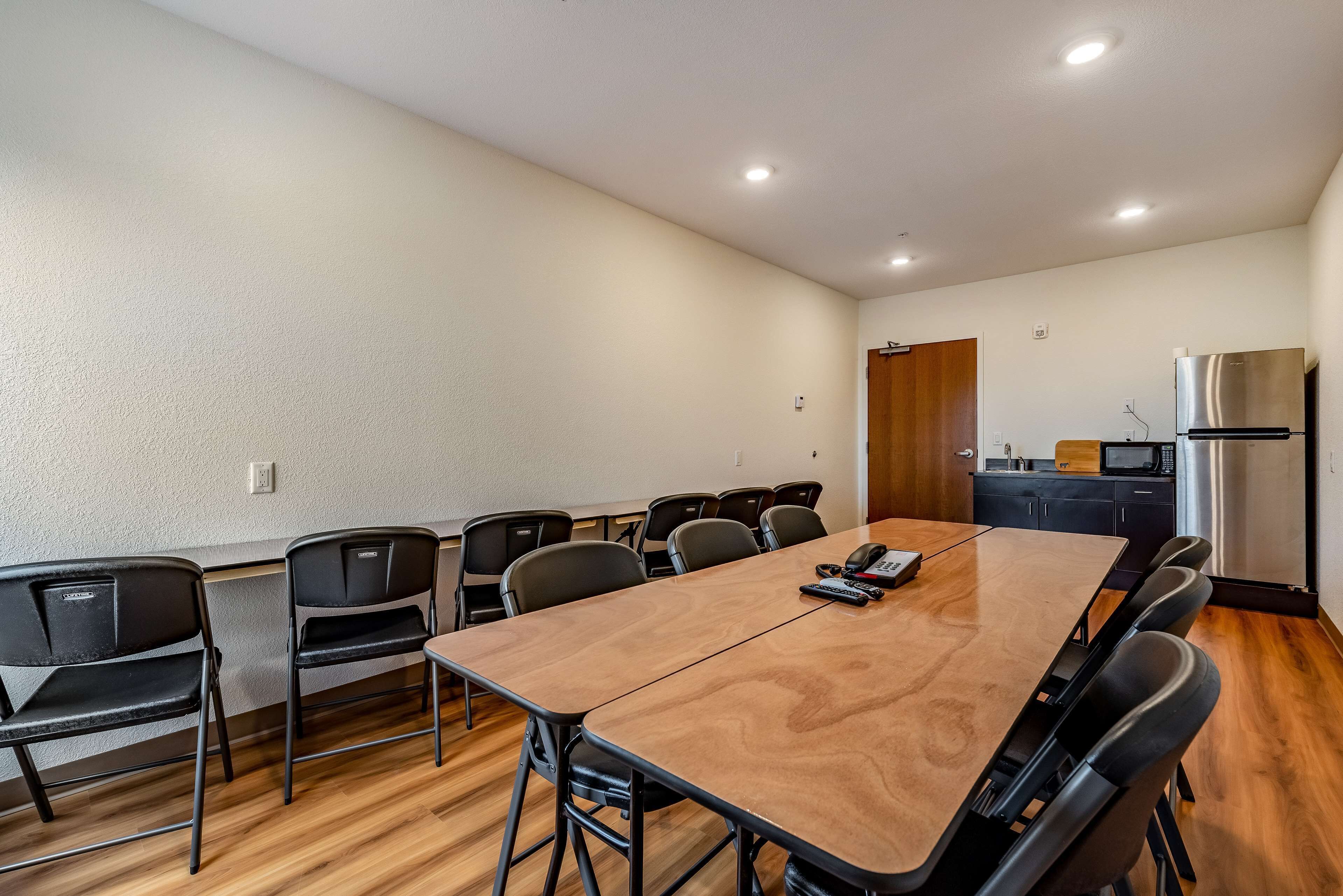 A conference room with a long table, black chairs, a wooden floor, a kitchen area with a refrigerator and microwave, and a door.