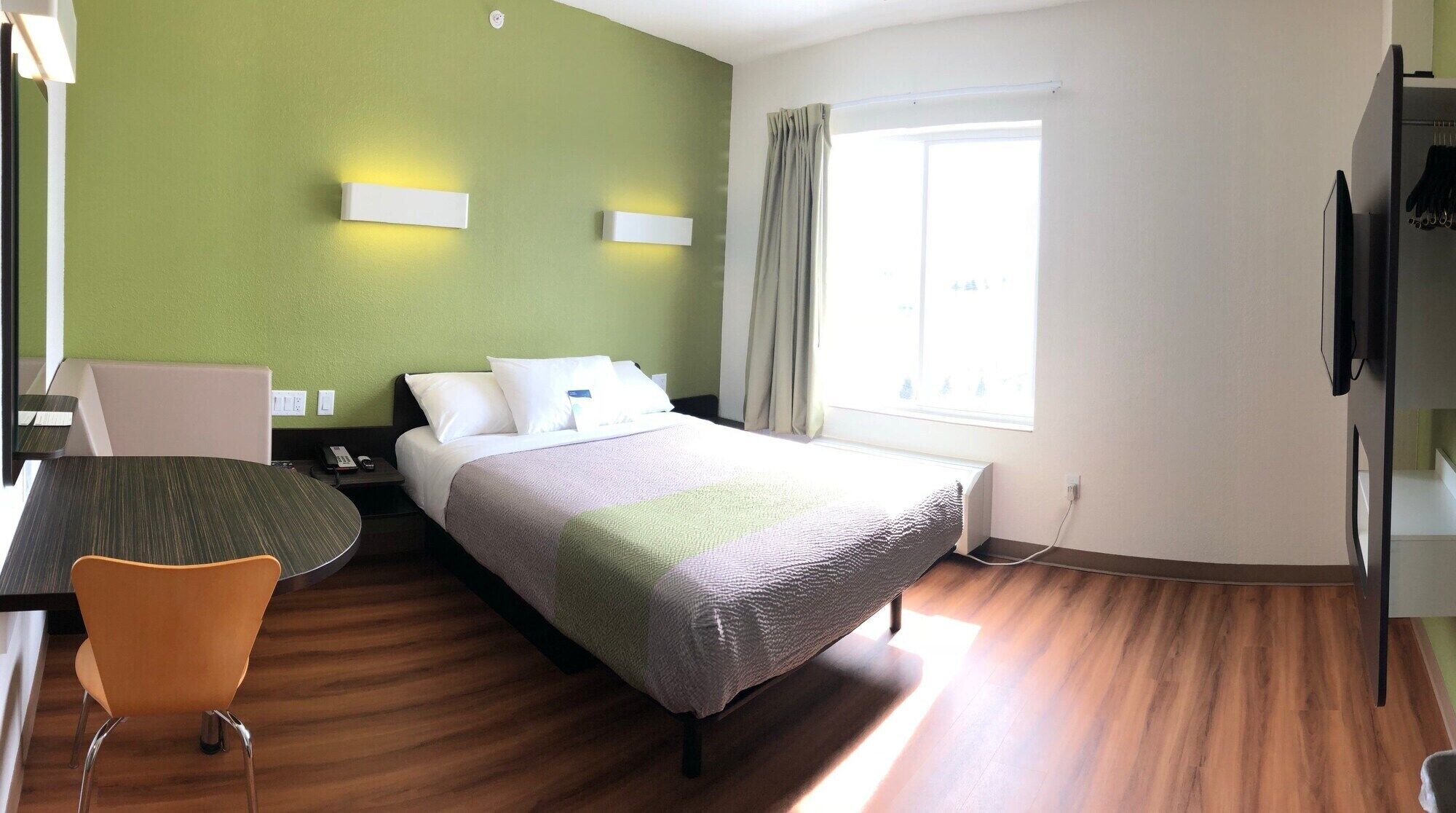 Et hotelværelse med seng, skrivebord, stol, fjernsyn og et vindue med gardiner.