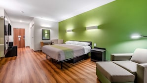 A modern hotel room with a wooden floor, a bed with a gray and white bedspread, a green accent wall, a desk with a chair, and a small seating area with a sofa.