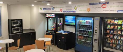 A modern kitchenette with a microwave, a coffee machine, and a small refrigerator. There are two tables with chairs and a vending machine with snacks.