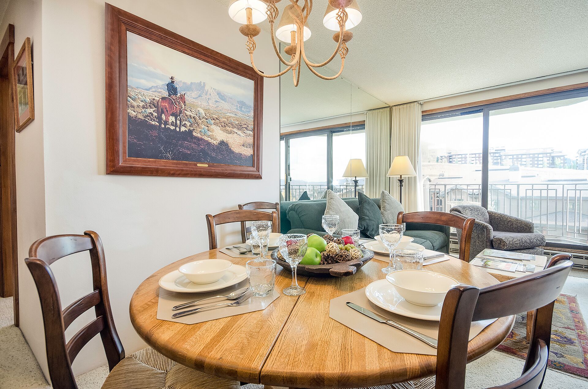 Un coin repas avec une table ronde en bois, dressée pour quatre personnes, entourée de chaises. Un tableau représentant un cow-boy à cheval est accroché au mur.