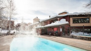 Un área de piscina con un jacuzzi, bancos y un edificio con techo nevado.