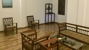 Une pièce avec un plancher en bois, un canapé, deux fauteuils, une petite table avec une théière et une étagère fixée au mur.
